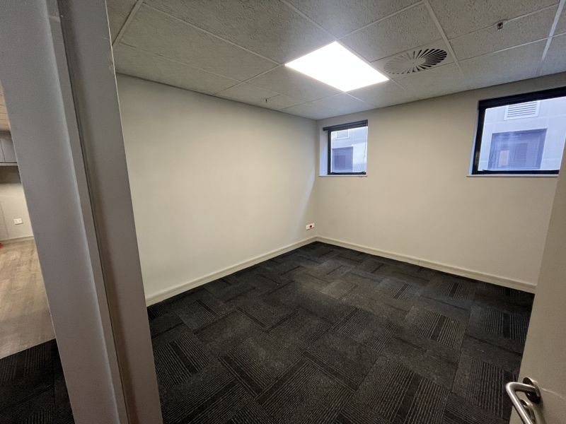 A clean, empty office space featuring neutral walls, dark carpet tiles, a suspended ceiling with recessed lighting, and air conditioning ventilation.