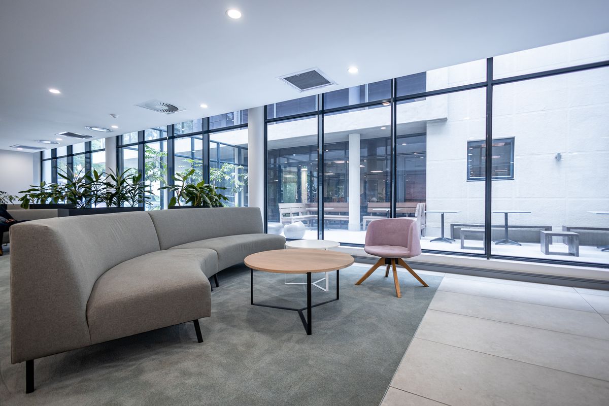 Modern office lounge area featuring floor-to-ceiling glass windows, recessed lighting, and a combination of tiled and carpeted flooring.