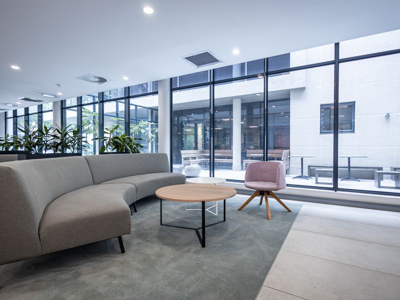 Modern office lounge area featuring floor-to-ceiling glass windows, recessed lighting, and a combination of tiled and carpeted flooring.