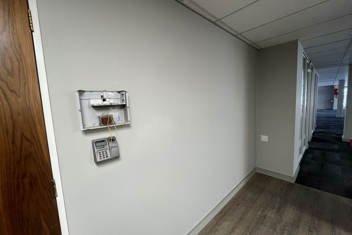 A hallway in an office space featuring a suspended ceiling, wood-look flooring, and an exposed alarm system control panel on the wall.