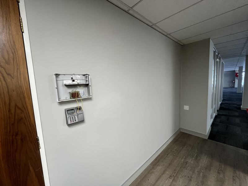 A hallway in an office space featuring a suspended ceiling, wood-look flooring, and an exposed alarm system control panel on the wall.