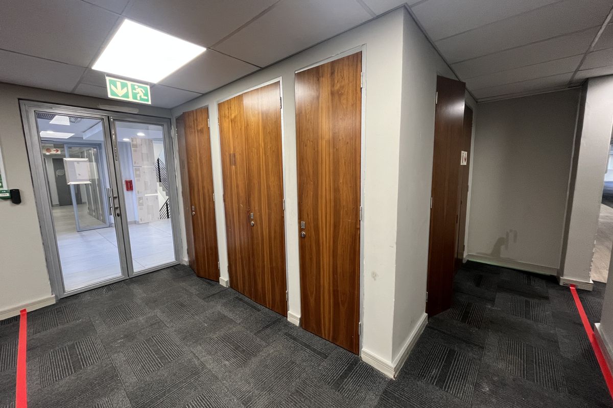 A functional office hallway featuring carpeted flooring, suspended ceiling with recessed lighting, and multiple wooden veneer doors leading to private office spaces.