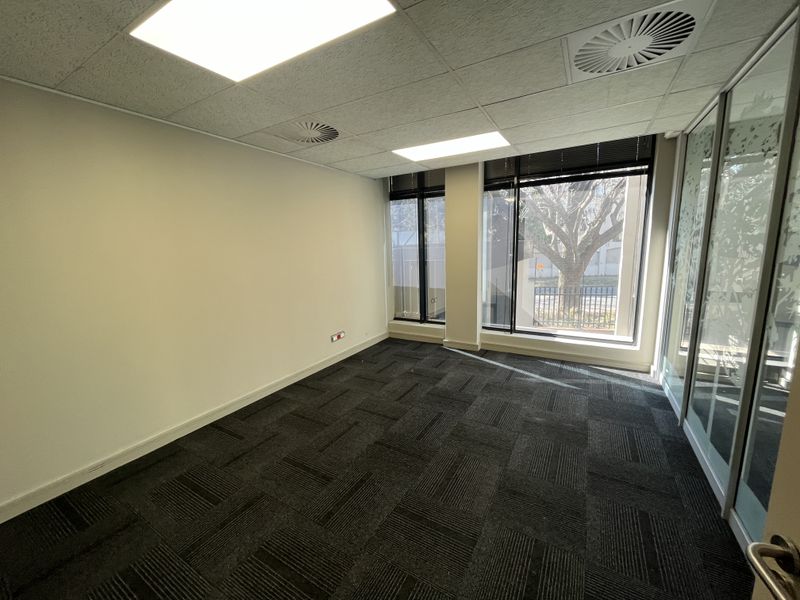 A bright, empty office space featuring modern carpet tiles, suspended ceiling with recessed lighting, and large windows providing natural light and a glass partition.