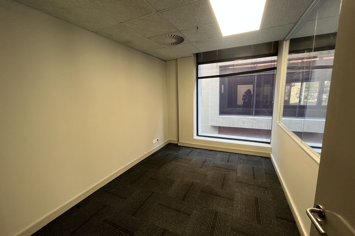 A vacant office space featuring carpet tiles, a suspended ceiling with recessed lighting, and a large window with a glass partition wall.