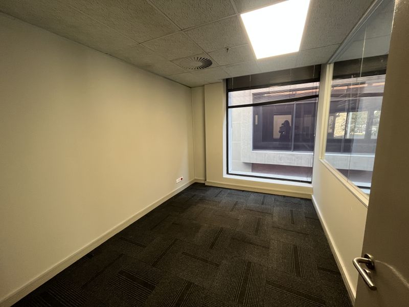 A vacant office space featuring carpet tiles, a suspended ceiling with recessed lighting, and a large window with a glass partition wall.