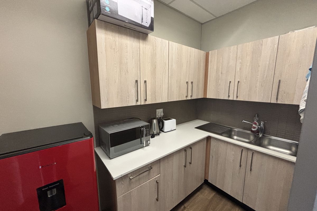 A compact office kitchenette featuring light wood-grain cabinetry, a stainless steel sink, and a tiled backsplash.