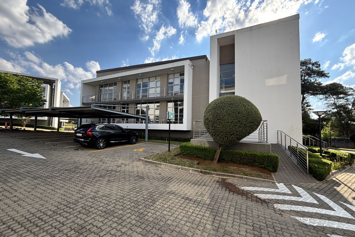 Modern multi-story office building exterior featuring a paved parking area, covered parking bays, and well-maintained landscaped grounds.