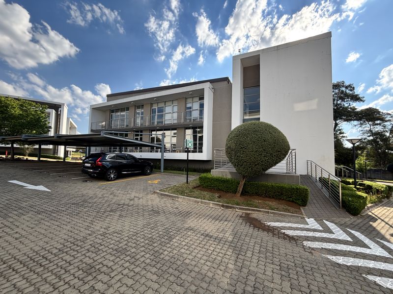 Modern multi-story office building exterior featuring a paved parking area, covered parking bays, and well-maintained landscaped grounds.