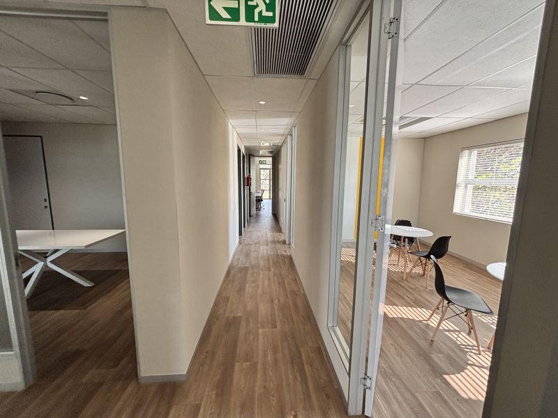 A bright office hallway featuring laminate flooring, glass partition walls, and integrated ceiling air conditioning vents.