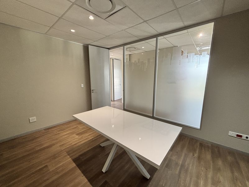 A modern office space featuring a glass partition wall with frosted detailing, suspended ceiling with recessed lighting, and durable laminate flooring.