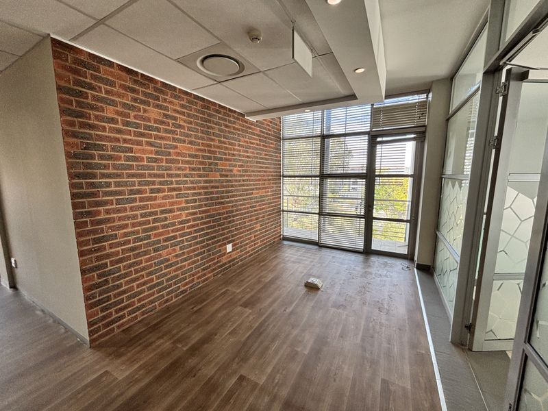 A bright, modern office space featuring an exposed brick feature wall, wood-look laminate flooring, and large windows with integrated blinds.