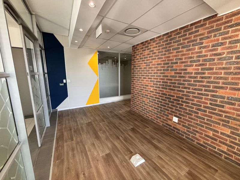 A modern, empty office space featuring an exposed brick feature wall, laminate flooring, glass partitions, and a suspended ceiling with recessed lighting.