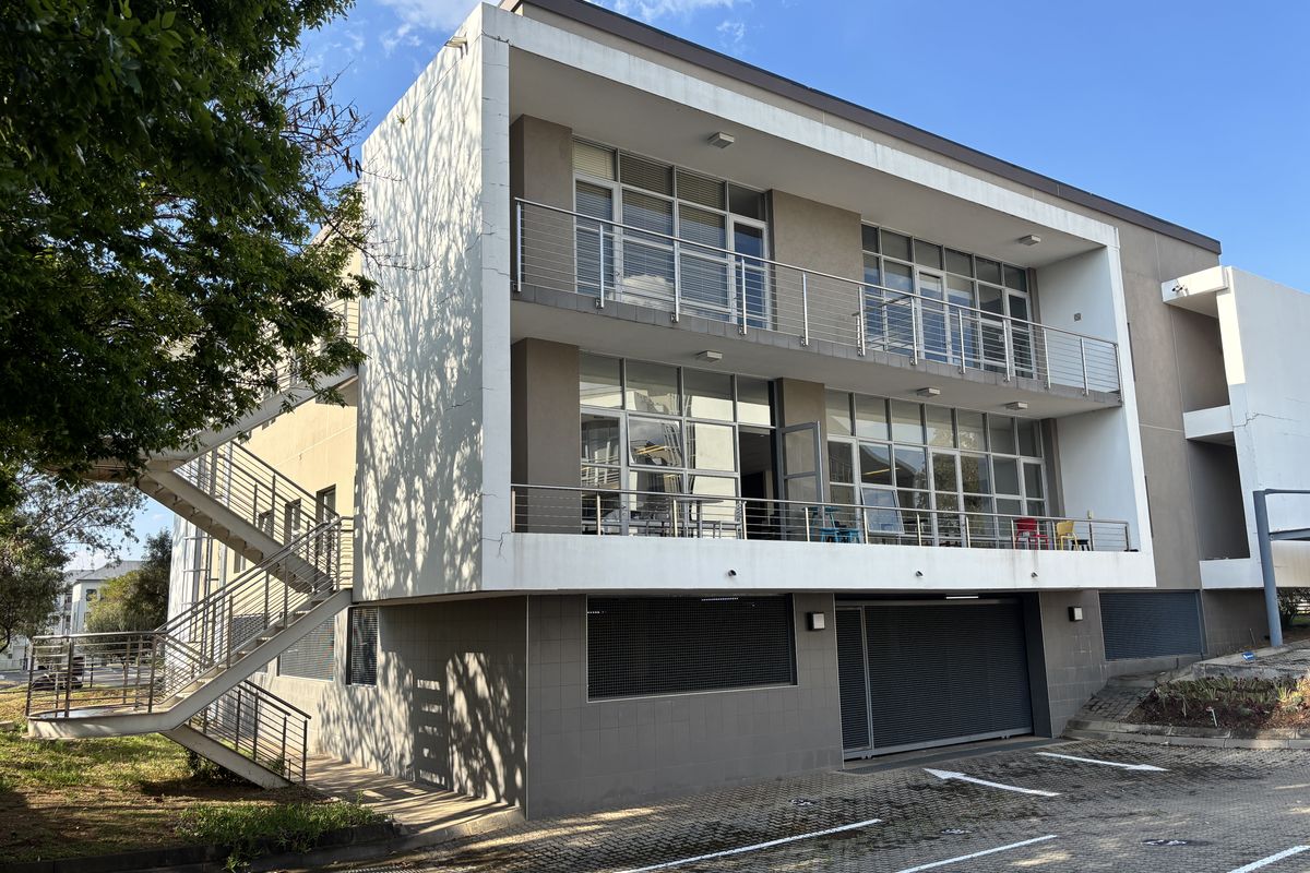 Modern multi-story office building featuring external staircases, private balconies with stainless steel railings, large glass windows, and a paved parking area.