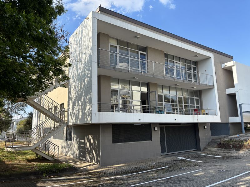 Modern multi-story office building featuring external staircases, private balconies with stainless steel railings, large glass windows, and a paved parking area.