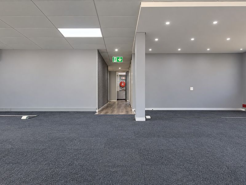 A spacious, modern open-plan office area featuring neutral grey walls, carpeted flooring, recessed lighting, and a clean, professional aesthetic.