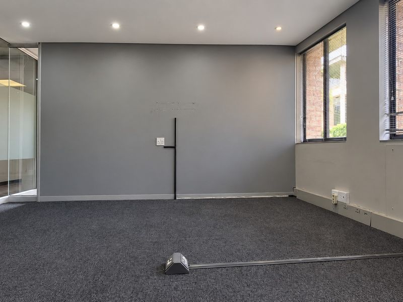 A vacant office space featuring grey carpeting, recessed lighting, glass partitioning, and large windows with venetian blinds, suitable for a professional workspace.