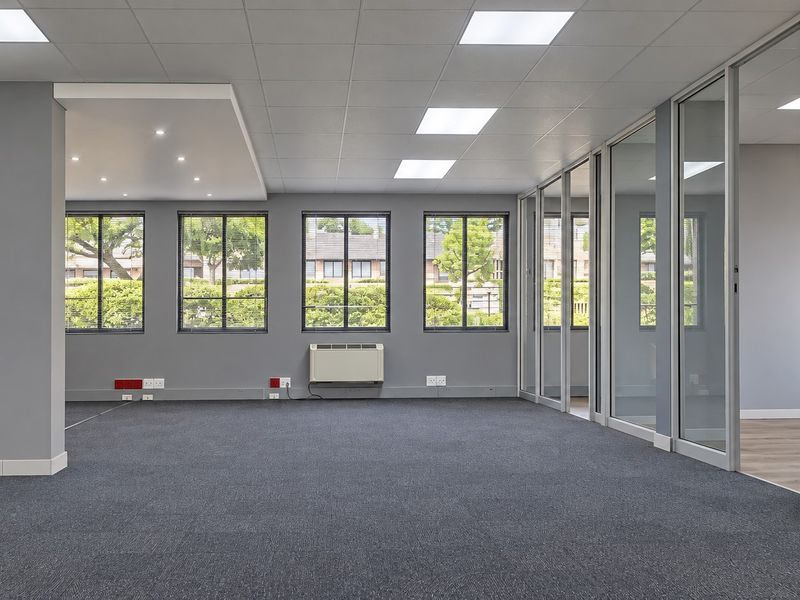A spacious, modern open-plan office area featuring glass partitions, carpeted flooring, recessed lighting, and large windows providing ample natural light.