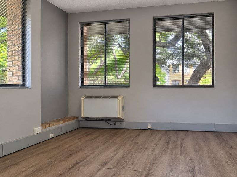 A bright, empty office space featuring wood-look laminate flooring, large windows with venetian blinds, and a wall-mounted air conditioning unit.