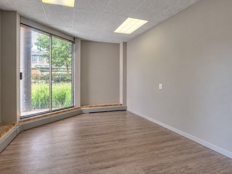 A bright, empty office space featuring wood-look flooring, a large window with blinds, and a suspended ceiling with recessed lighting.