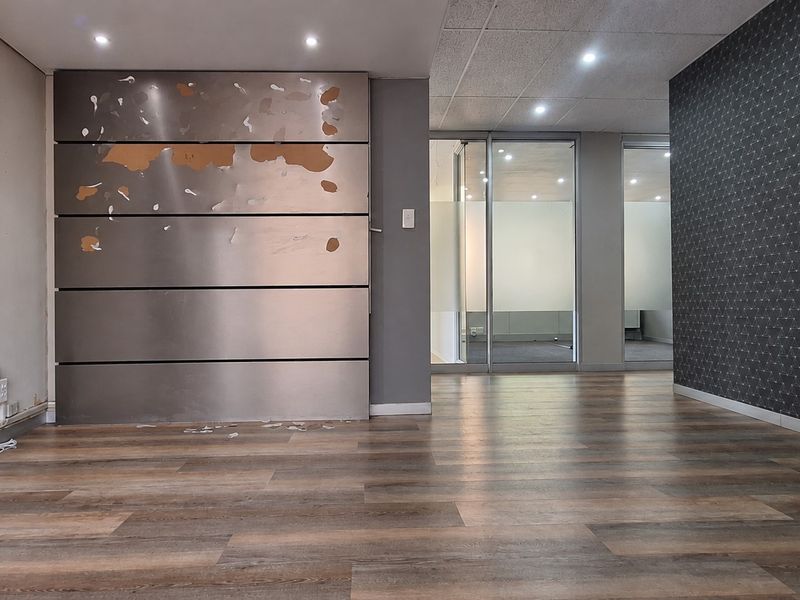 Empty office space featuring wood-look laminate flooring, recessed lighting, and glass partitions, with a damaged metal feature wall requiring repair.