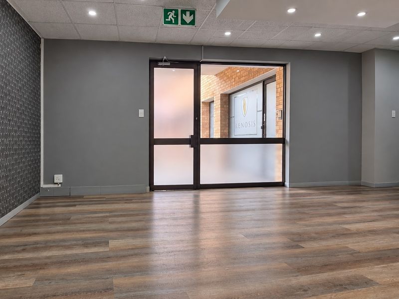 A bright, empty office space featuring modern laminate flooring, a suspended ceiling with recessed lighting, and a frosted glass entrance door.