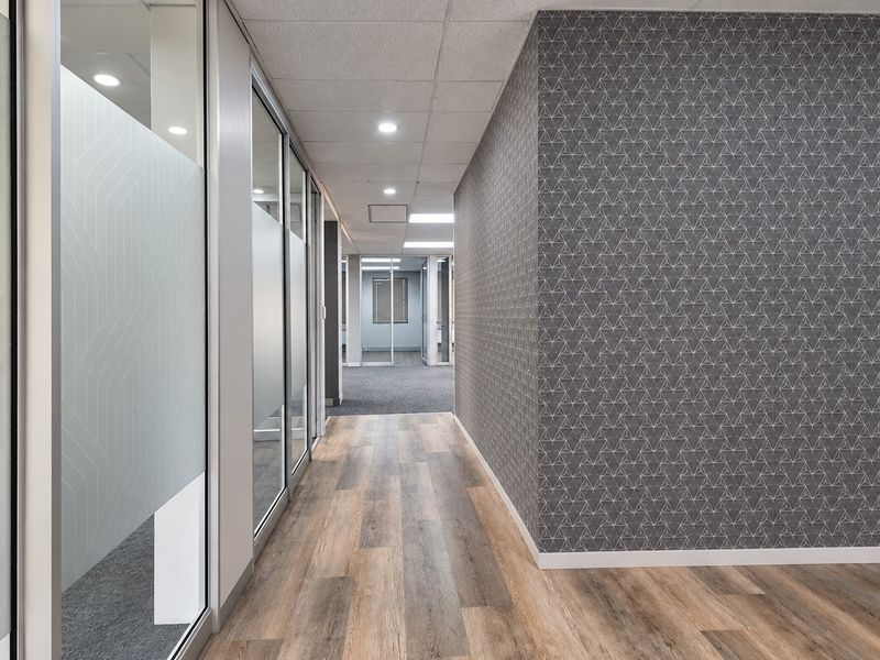 Modern office hallway featuring glass partitions, wood-look vinyl flooring, and a feature wall with geometric patterned wallpaper.