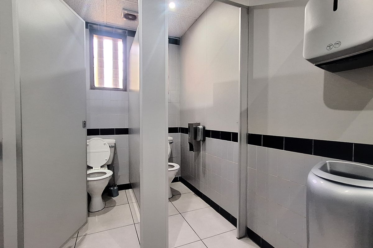 A functional commercial office bathroom featuring tiled walls and flooring, partitioned toilet stalls, and integrated wall-mounted dispensers.