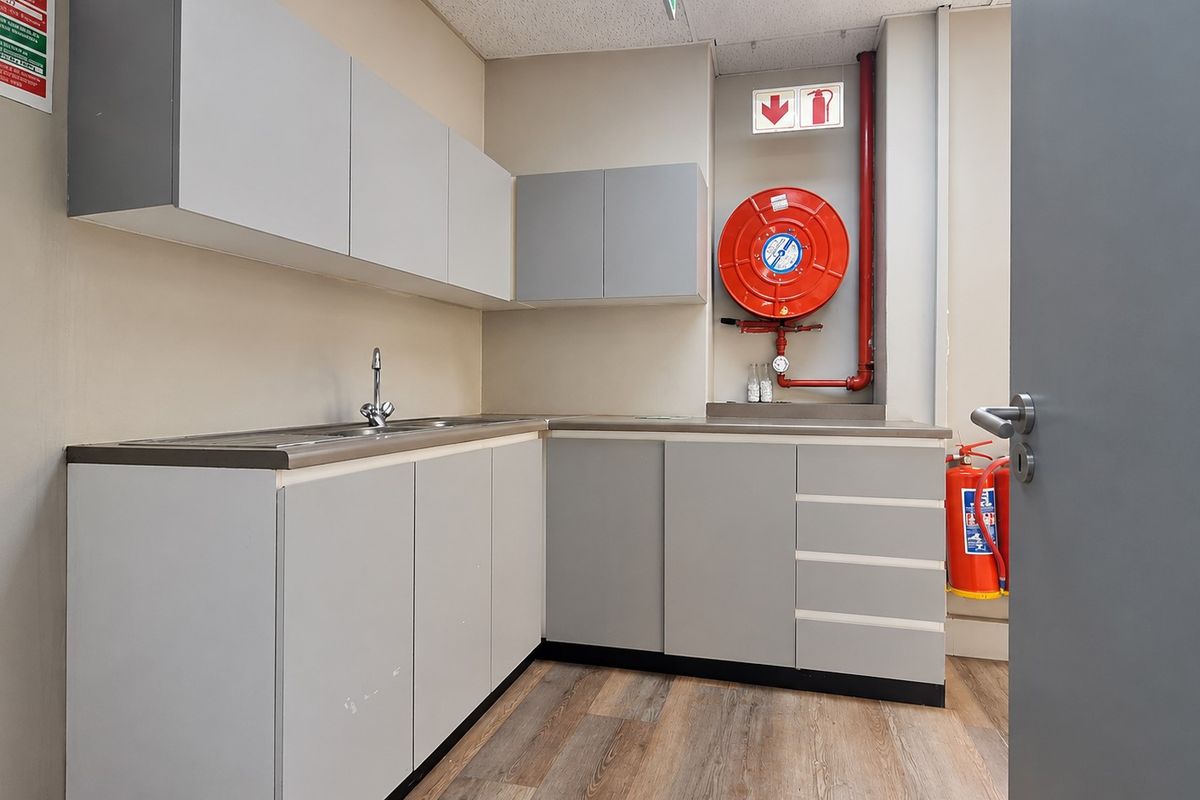 A functional office kitchenette featuring grey built-in cabinetry, a stainless steel sink, and essential fire safety equipment including a hose reel and extinguisher.
