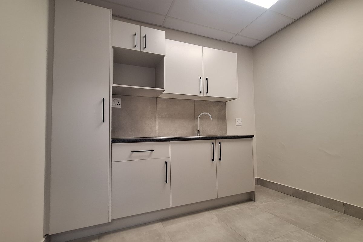 A clean, modern office kitchenette featuring neutral cabinetry, a sink, tiled backsplash, and durable tiled flooring.