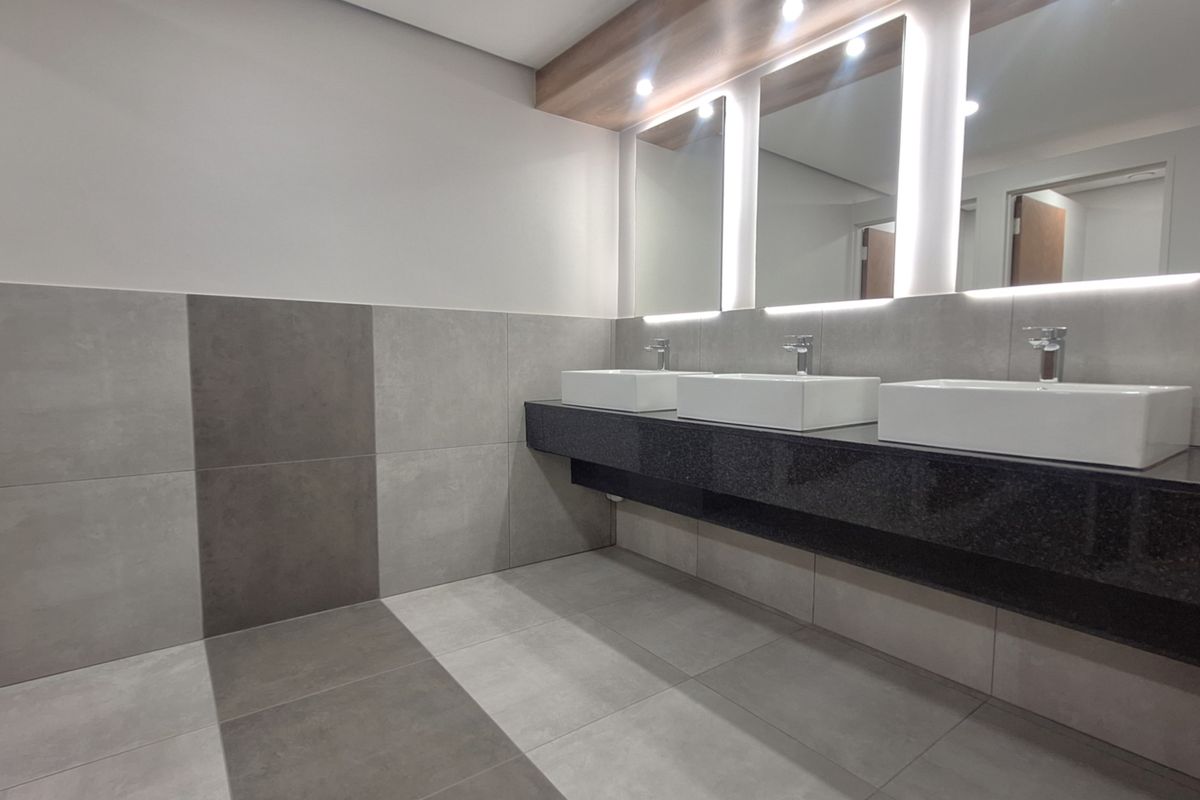 Modern office bathroom featuring a triple vanity with vessel sinks, granite countertop, backlit mirrors, and contemporary large-format grey tiling.