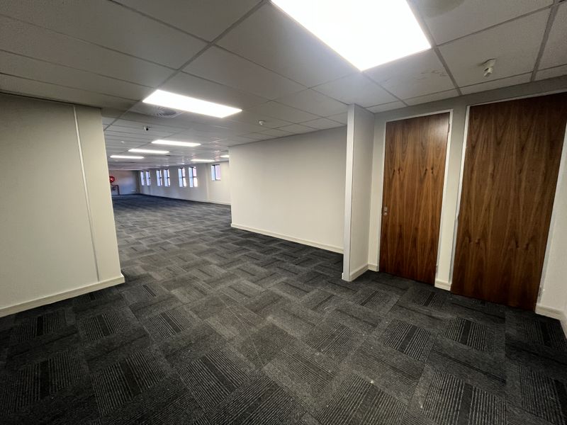 A spacious, empty open-plan office area featuring carpet tiles, suspended ceilings with recessed lighting, and two wooden veneer doors.
