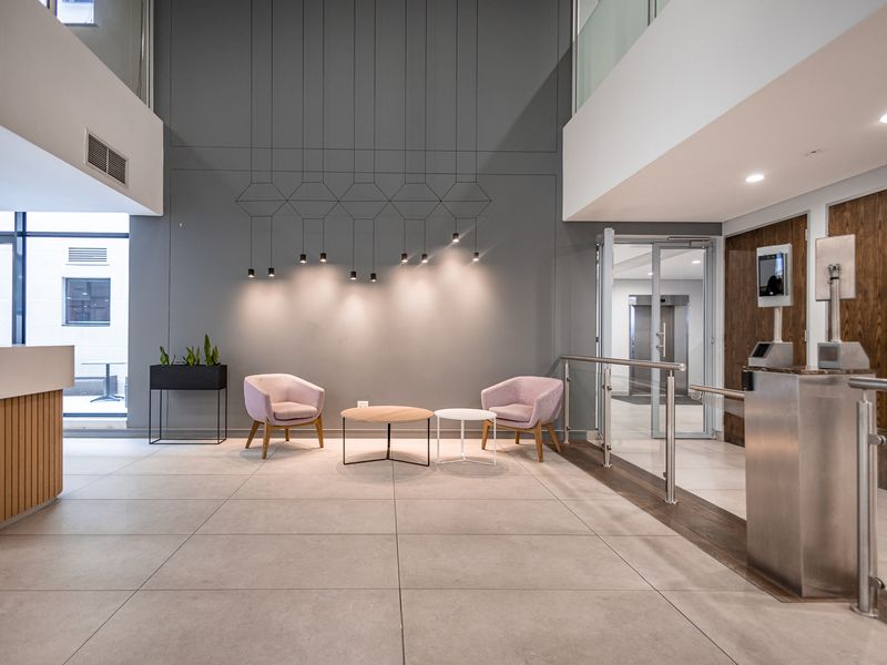 Modern office building lobby featuring a reception area, security turnstiles, high ceilings, and contemporary lighting fixtures with polished tiled flooring.