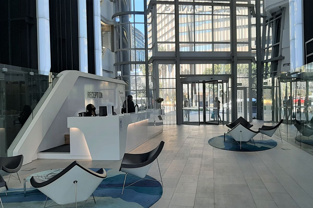 Modern office building entrance lobby featuring a sleek reception desk, expansive floor-to-ceiling glass facade, and high-quality tiled flooring.