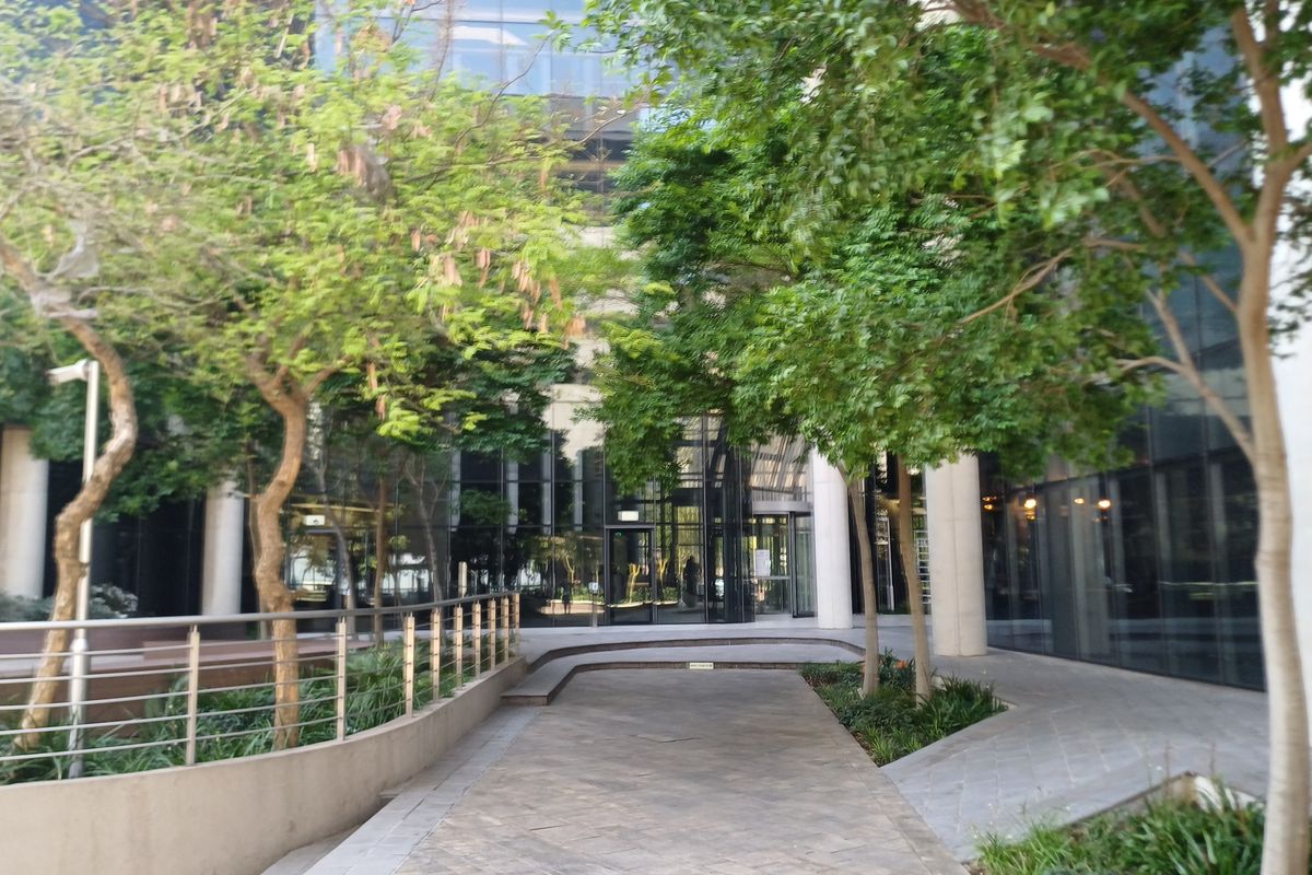 Modern office building entrance featuring a glass facade, paved pedestrian walkway, and landscaped garden areas.