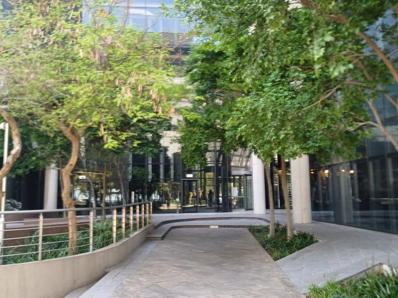 Modern office building entrance featuring a glass facade, paved pedestrian walkway, and landscaped garden areas.