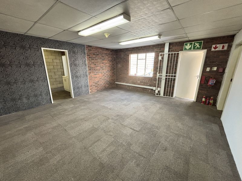 An empty office space featuring exposed brick walls, suspended ceiling with fluorescent lighting, and carpeted flooring, equipped with security gate and fire safety equipment.