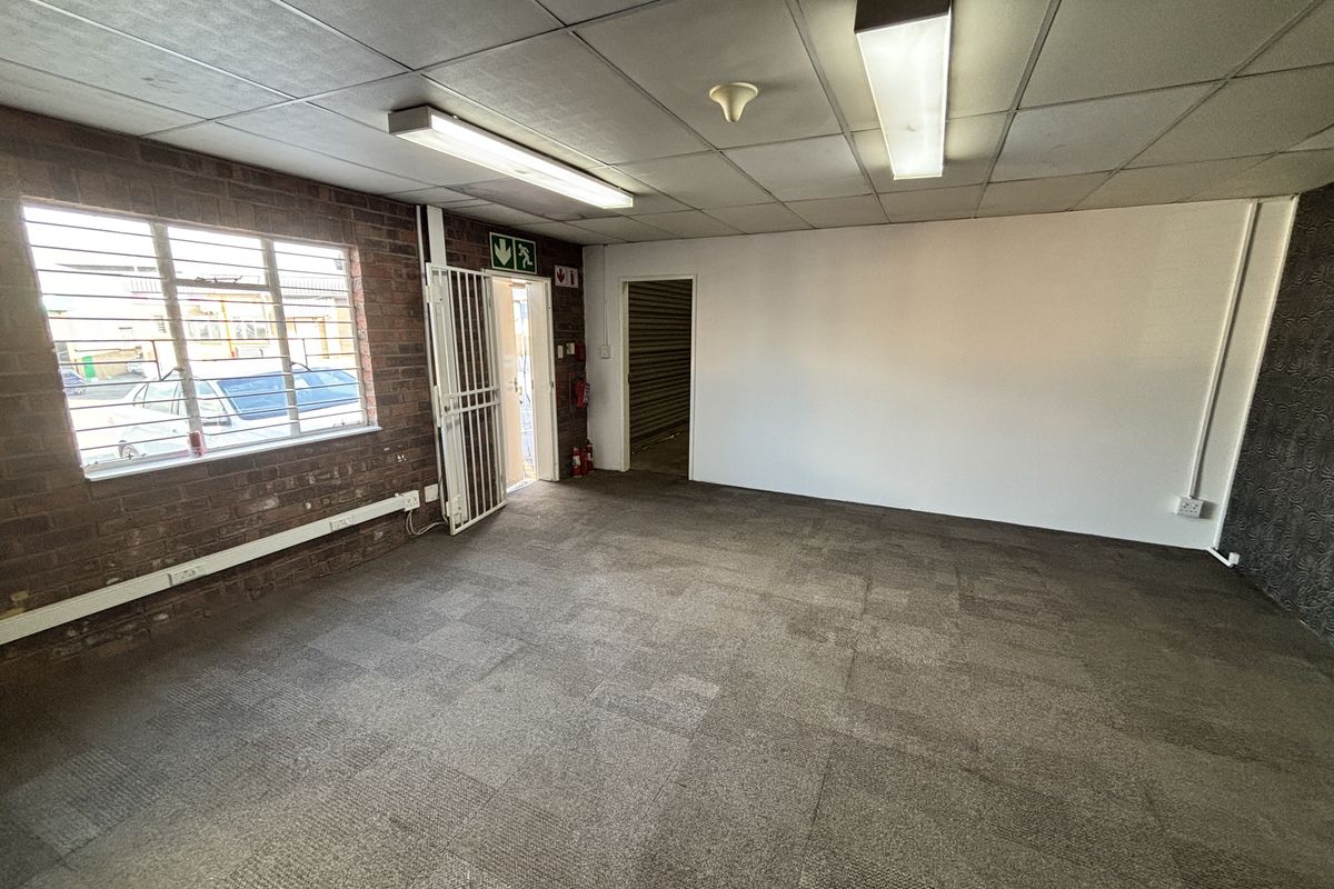 An empty industrial office space featuring exposed brickwork, carpeted flooring, suspended ceiling with fluorescent lighting, and a secure metal entrance gate.