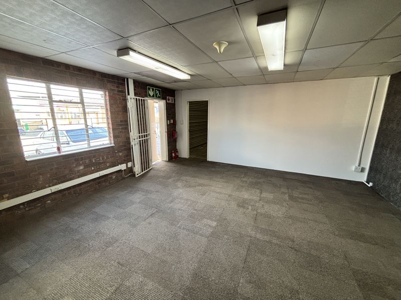 An empty industrial office space featuring exposed brickwork, carpeted flooring, suspended ceiling with fluorescent lighting, and a secure metal entrance gate.