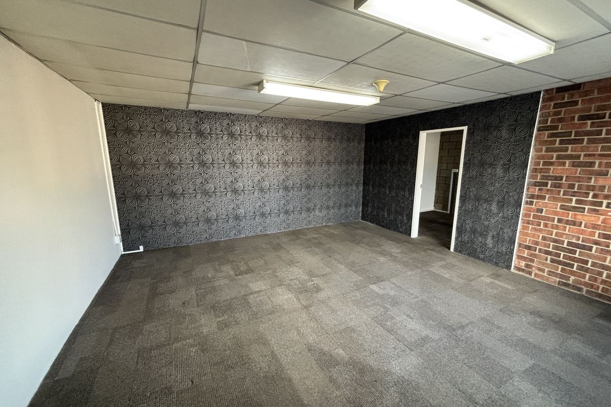 An empty office space featuring a suspended ceiling, fluorescent lighting, exposed brick accents, and textured wallpaper with carpeted flooring.