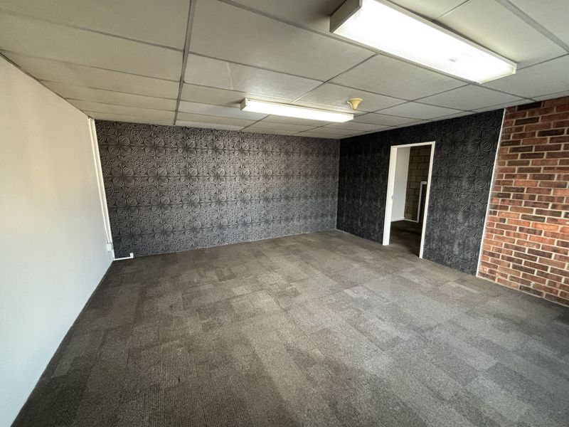 An empty office space featuring a suspended ceiling, fluorescent lighting, exposed brick accents, and textured wallpaper with carpeted flooring.