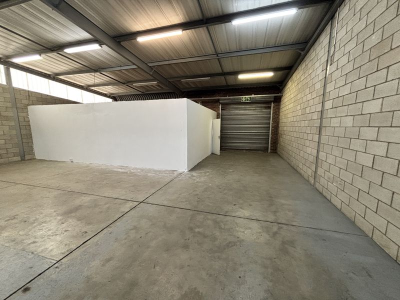 Spacious industrial garage or warehouse unit featuring high ceilings with exposed steel trusses, concrete block walls, and a secure roller shutter door.