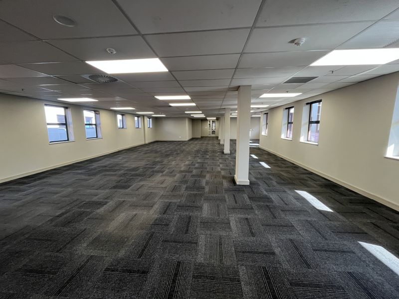 A spacious, open-plan office area featuring neutral walls, carpeted flooring, suspended ceilings with recessed lighting, and multiple windows providing natural light.