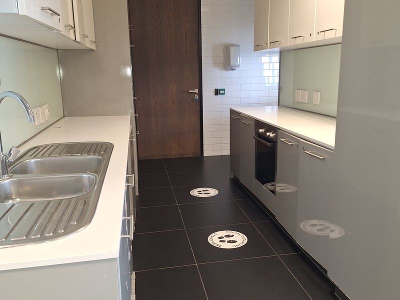 A clean, modern office kitchenette featuring grey cabinetry, stainless steel sink, built-in oven, and dark tiled flooring.