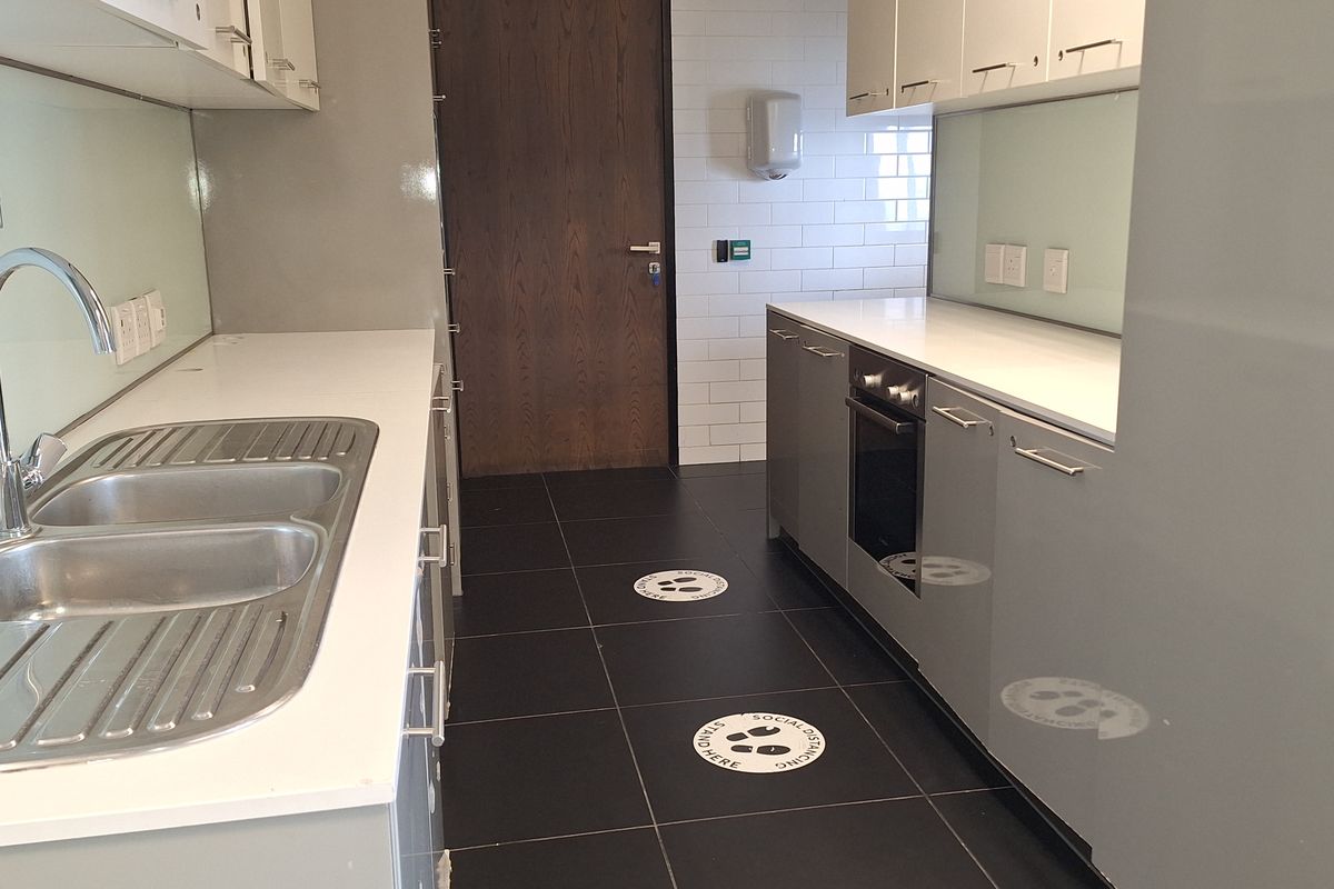 A clean, modern office kitchenette featuring grey cabinetry, stainless steel sink, built-in oven, and dark tiled flooring.