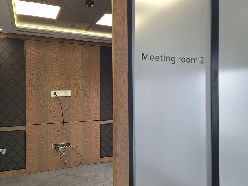 A modern office meeting room featuring wood veneer wall paneling, recessed ceiling lights, and a frosted glass entrance door.