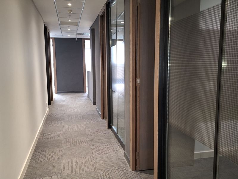 A modern office hallway featuring carpet tiles, recessed ceiling lighting, and glass partition walls with frosted privacy detailing.