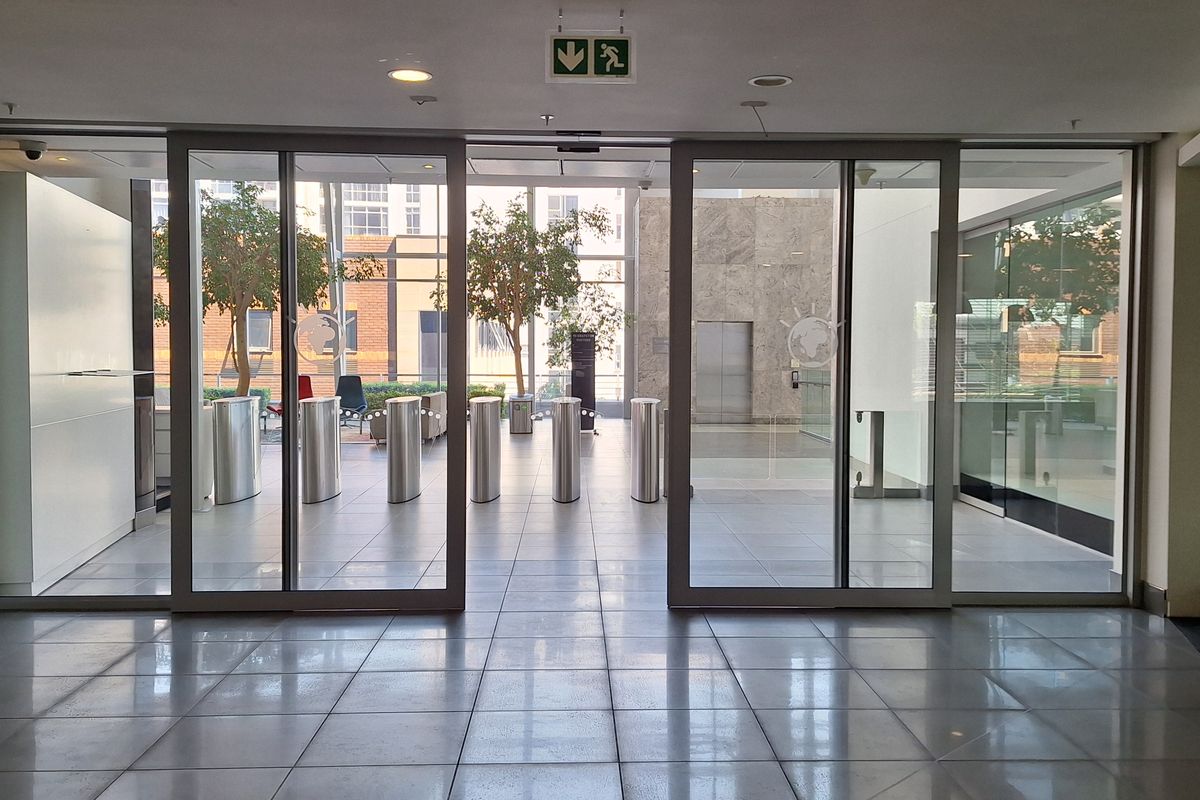 Modern office building entrance lobby featuring glass sliding doors, security turnstiles, and polished tiled flooring with recessed lighting.