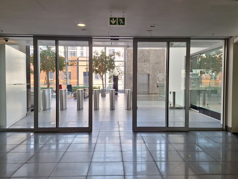 Modern office building entrance lobby featuring glass sliding doors, security turnstiles, and polished tiled flooring with recessed lighting.