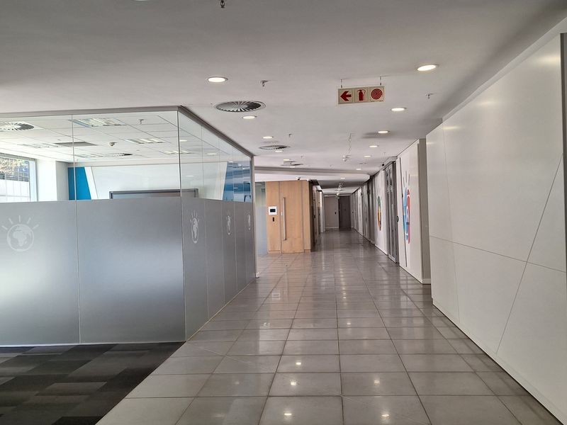 A spacious, well-lit office hallway featuring polished ceramic floor tiles, glass partitions, and integrated ceiling infrastructure including fire safety systems.