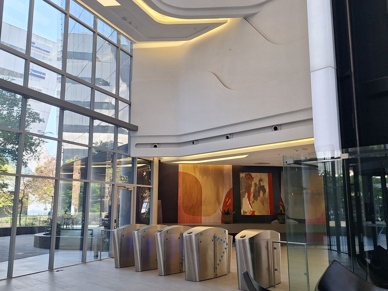 Modern office building lobby featuring a double-volume glass facade, integrated security turnstiles, and sophisticated architectural ceiling lighting.